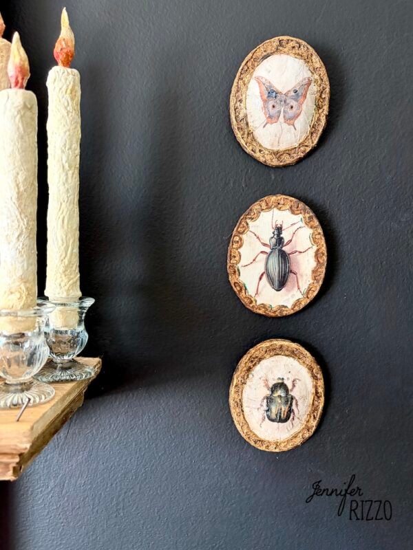 Three decorative wall plaques featuring insect and butterfly illustrations are displayed vertically next to a shelf with lit candles against a dark wall.
