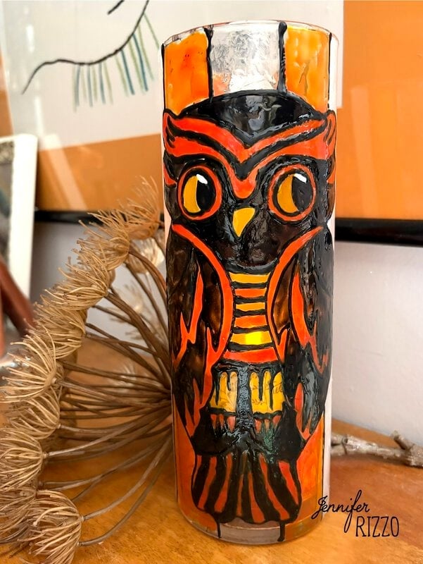 A clear glass with a painted orange and black owl design sits on a wooden surface next to dried foliage.