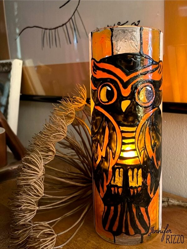 A glass candle holder with a painted owl design glows with an orange light. Decorative objects and wall art are visible in the background.