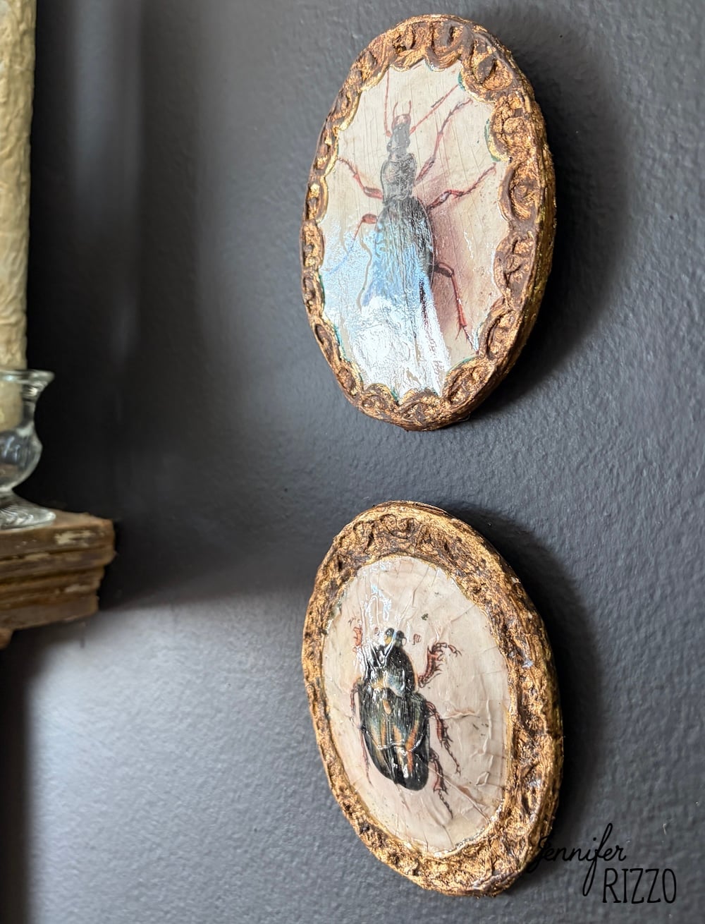 Two small, round plaques with painted beetles and textured gold borders hang on a dark gray wall.