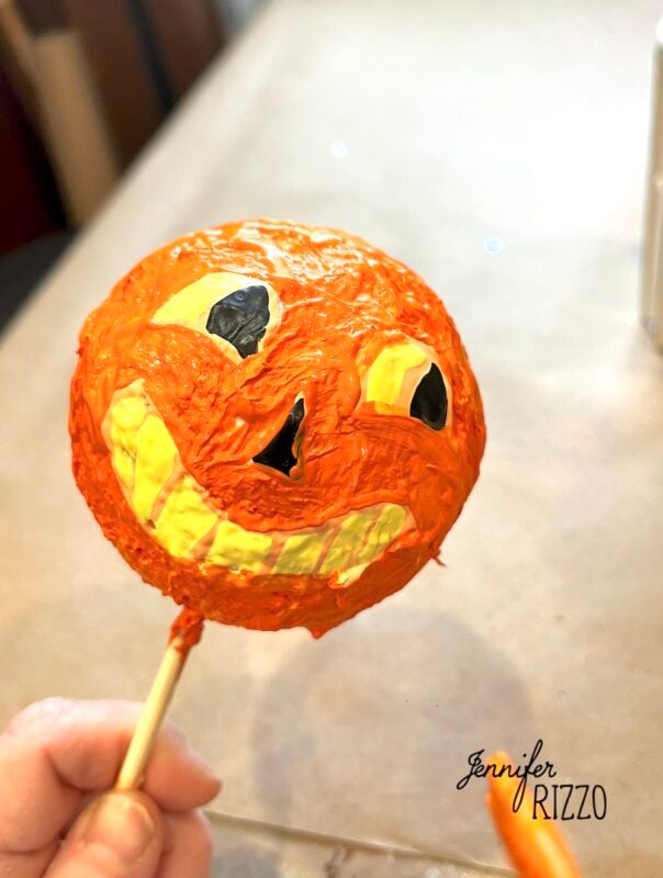 Hand holding a round orange craft with a painted yellow and black jack-o’-lantern face on a stick. “Jennifer Rizzo” is written in the bottom right corner.