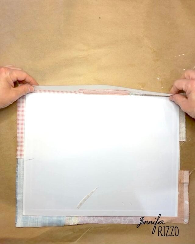 Two hands fold fabric over the edge of a white rectangular object on a brown work surface with “Jennifer Rizzo” text in the corner.