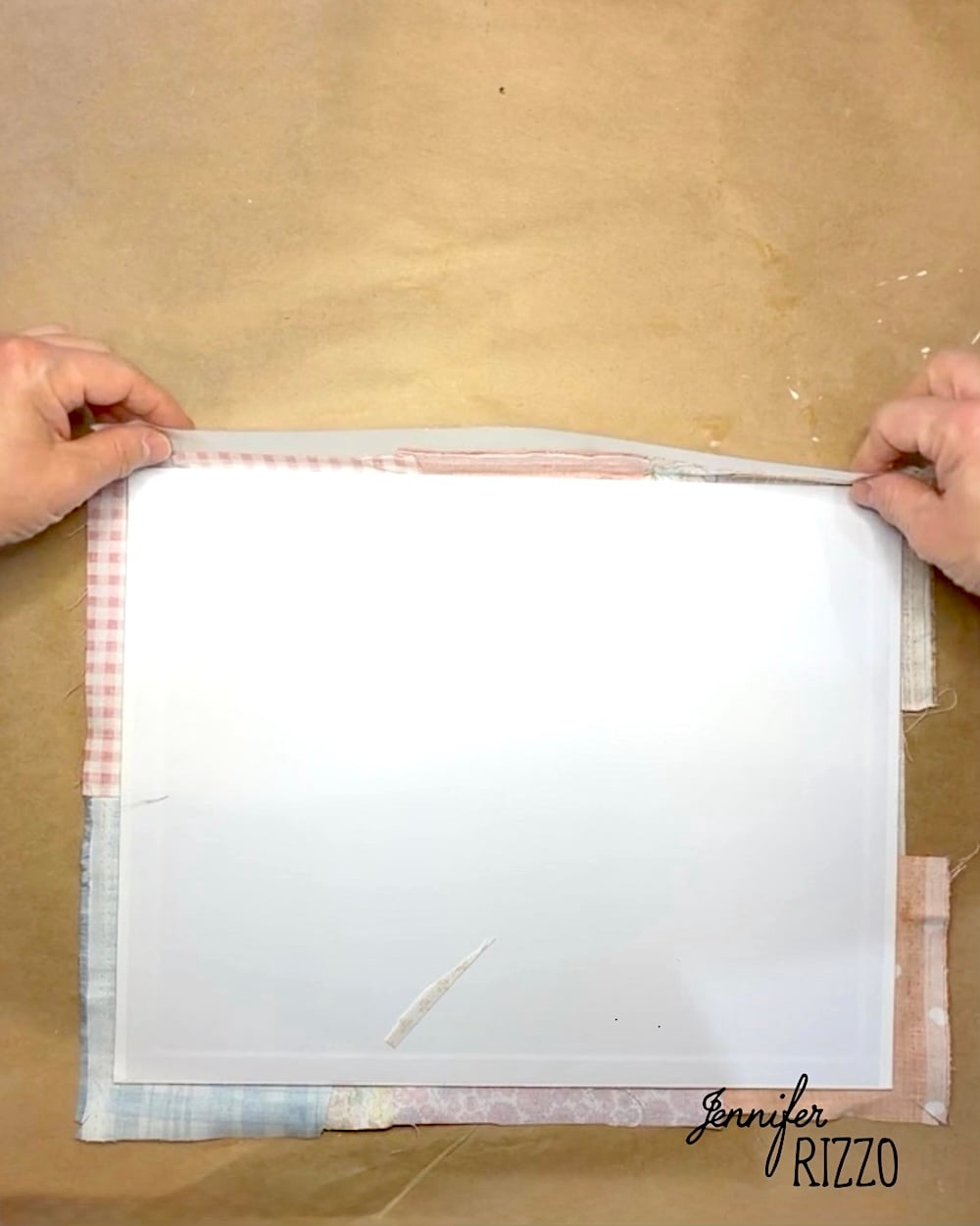Two hands fold fabric over the edge of a white rectangular object on a brown work surface with “Jennifer Rizzo” text in the corner.
