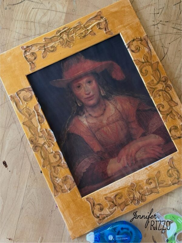 Framed portrait of a woman in a red outfit and hat on a wooden table, with decorative frame details and tape dispensers nearby.