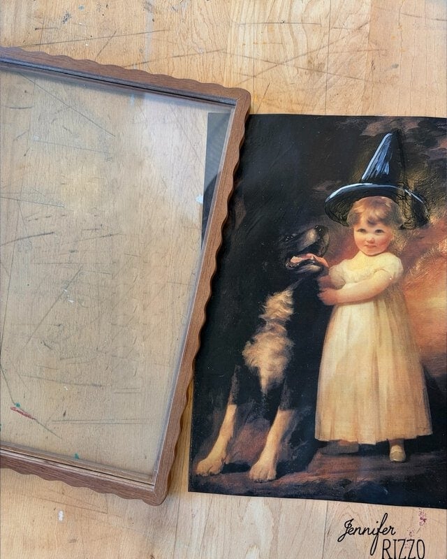 A framed glass and a vintage-style print of a child in a white dress with a dog; the child is wearing a black pointed hat.