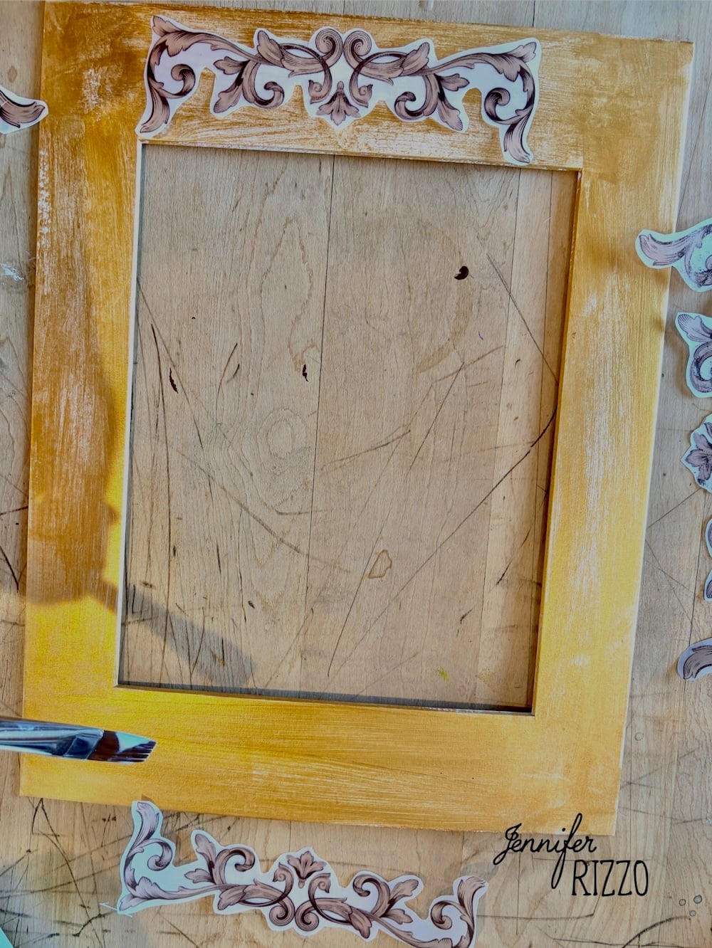 A wooden picture frame partially painted yellow lies on a scratched surface, surrounded by decorative corner pieces and a paintbrush. The name “Jennifer Rizzo” appears in the lower right corner.