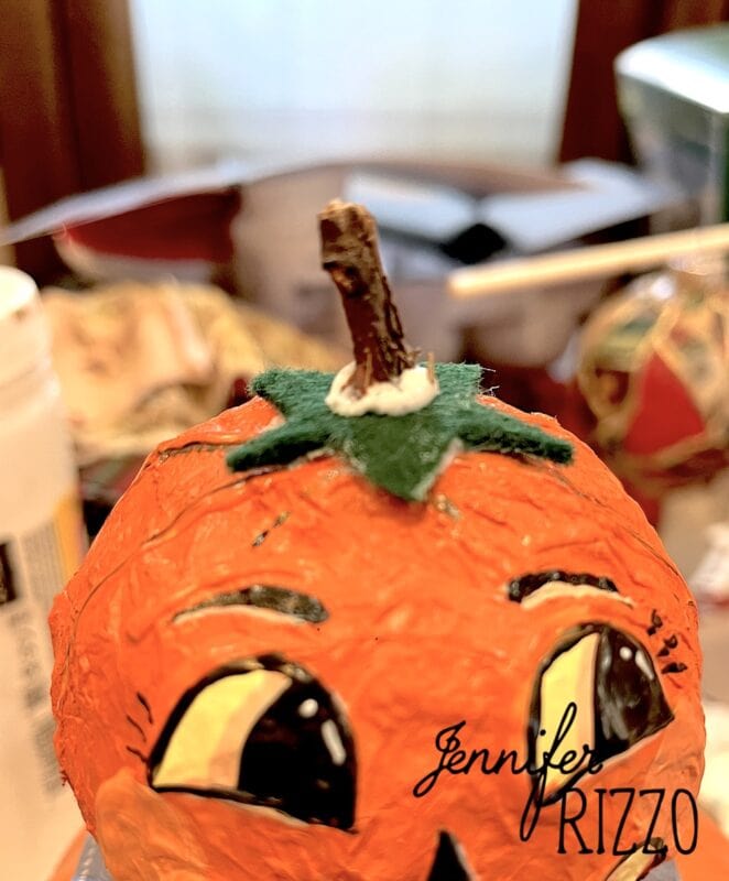 Close-up of a handmade pumpkin craft with painted eyes, a green felt stem, and a small stick on top. The name “Jennifer Rizzo” appears in the lower right corner.