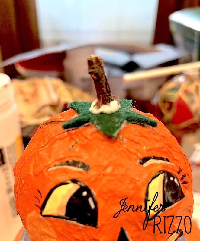 Close-up of a handmade pumpkin craft with painted eyes, a green felt stem, and a small stick on top. The name “Jennifer Rizzo” appears in the lower right corner.