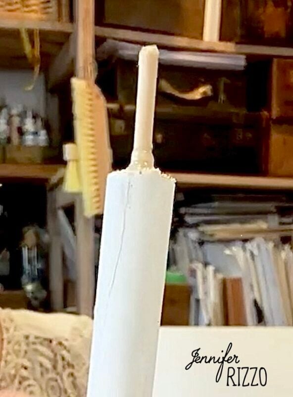 A close-up of a white cylindrical object, resembling one of the popular Spun Cotton Faux Candles with a thin stick protruding from the top, set against a background of shelves, books, and craft supplies.