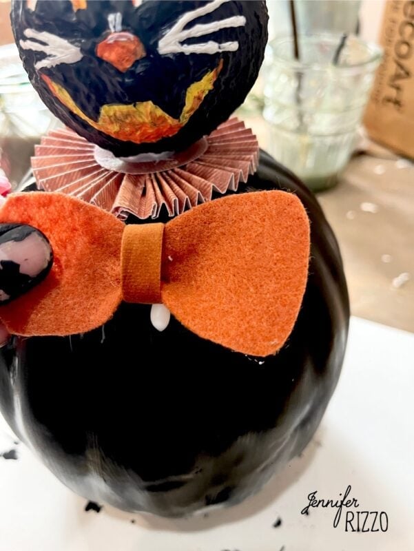 A close-up of a handmade black cat Halloween decoration with an orange felt bow tie and a ruffled paper collar.