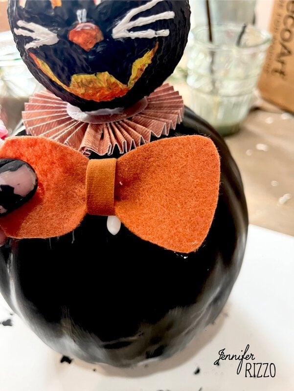 A close-up of a handmade black cat Halloween decoration with an orange felt bow tie and a ruffled paper collar.