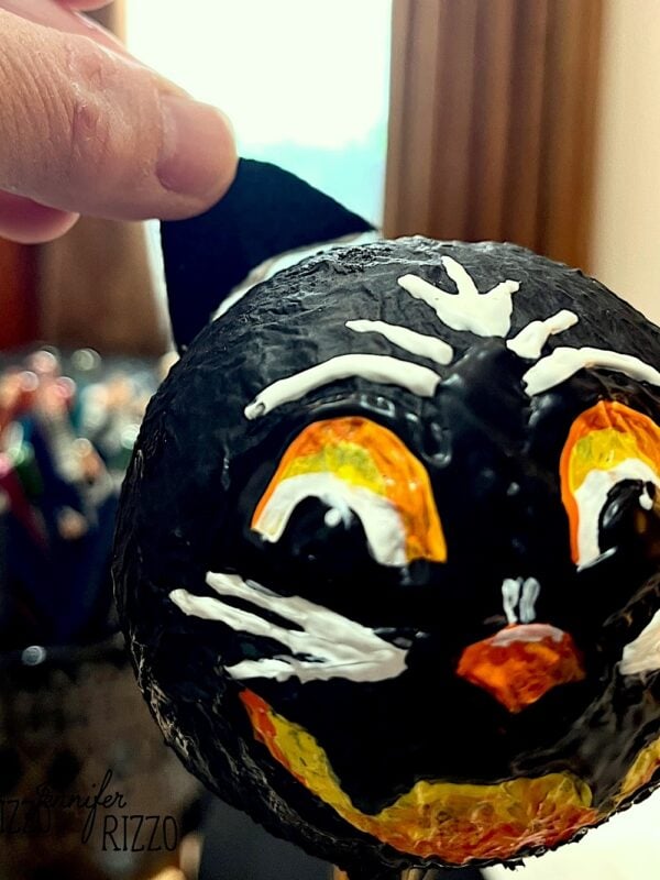 A hand holds a black, cat-shaped craft with painted white, orange, and yellow facial features.