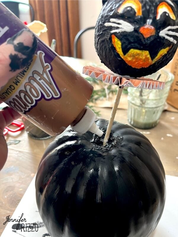 A hand applies craft glue to the top of a painted black pumpkin; a painted cat head decoration is attached to a stick above the pumpkin.