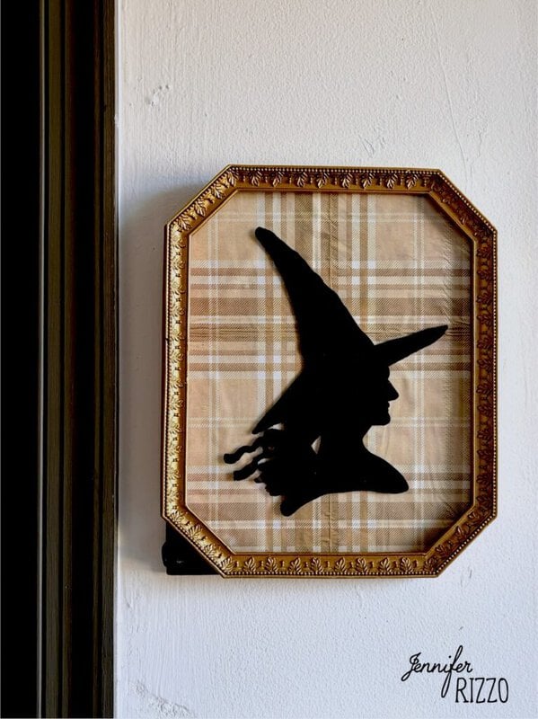 Framed silhouette of a witch with a pointed hat on a plaid background, hung on a white wall.