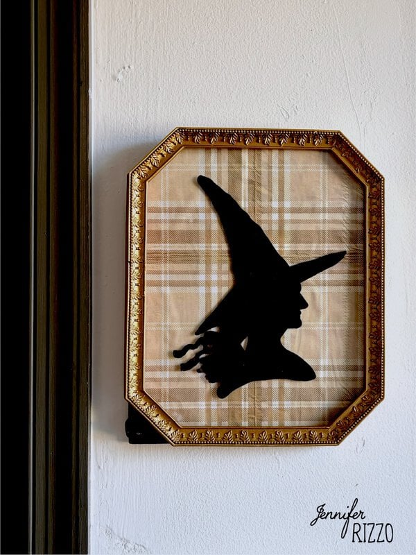 Framed silhouette of a witch with a pointed hat on a plaid background, hung on a white wall.