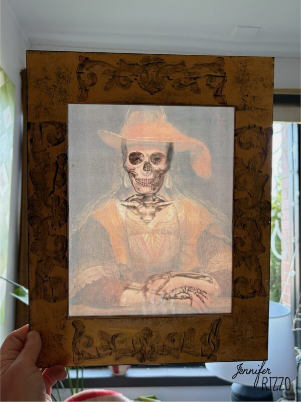 A hand holds a framed portrait of a woman wearing a hat; the face and hands are altered to appear as a skeleton.