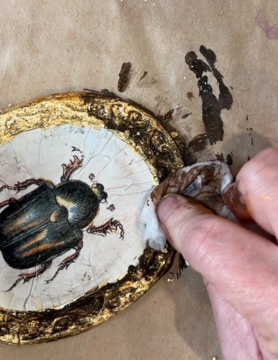 A hand wipes around an oval painting of a beetle with a gold ornate border on brown paper.