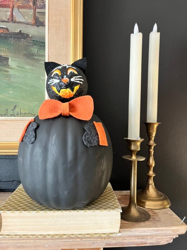 A black pumpkin decorated as a cat with an orange bow sits on a book next to two unlit candles in brass holders.