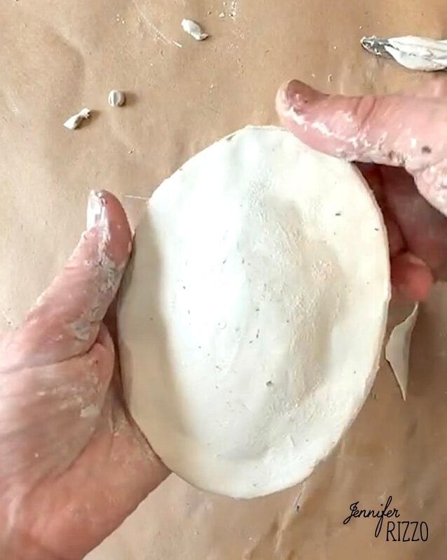 Two hands shaping a flat, oval piece of white clay on a brown work surface, with small clay scraps and the name “Jennifer Rizzo” visible in the corner.
