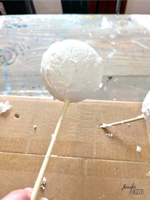 A hand holds a white, textured ball on a wooden stick over a table with craft materials.