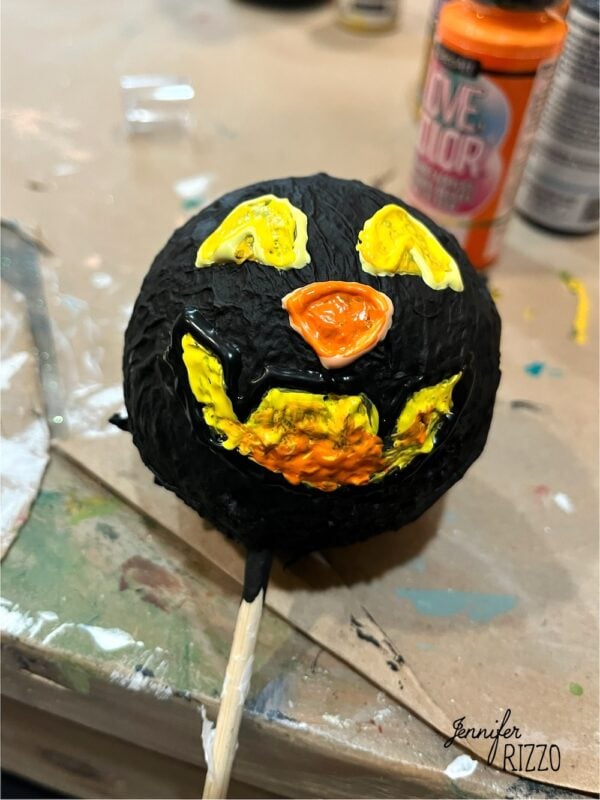 A round object painted black with yellow and orange facial features resembling a jack-o’-lantern, placed on a stick on a messy work surface.