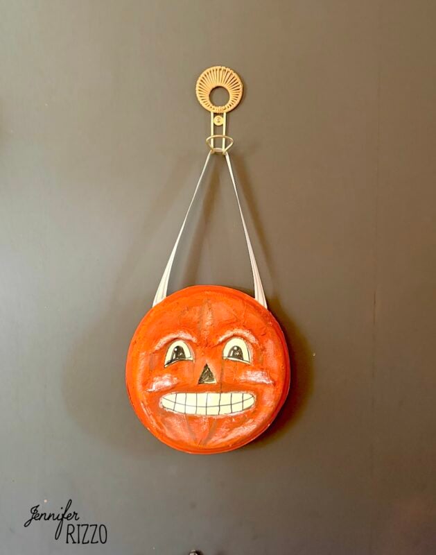 Round orange object with a painted jack-o’-lantern face, white strap, and metal hook hangs on a dark wall. “Jennifer Rizzo” is written in the lower left corner.