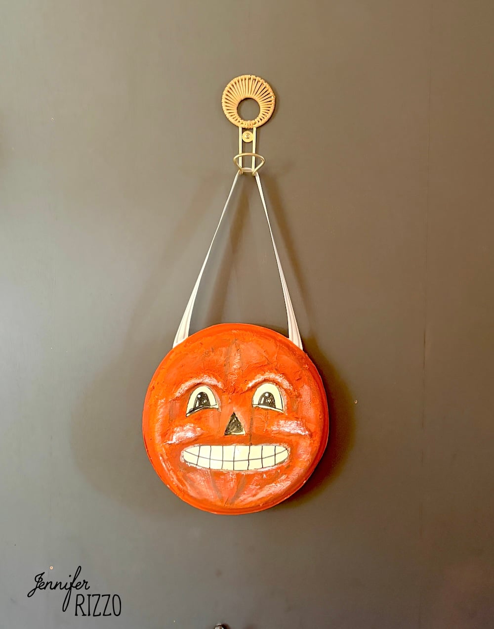 Round orange object with a painted jack-o’-lantern face, white strap, and metal hook hangs on a dark wall. “Jennifer Rizzo” is written in the lower left corner.