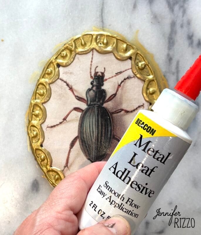 A hand holds a tube of metal leaf adhesive near an oval frame with a beetle illustration and a gold rim, on a marble surface.