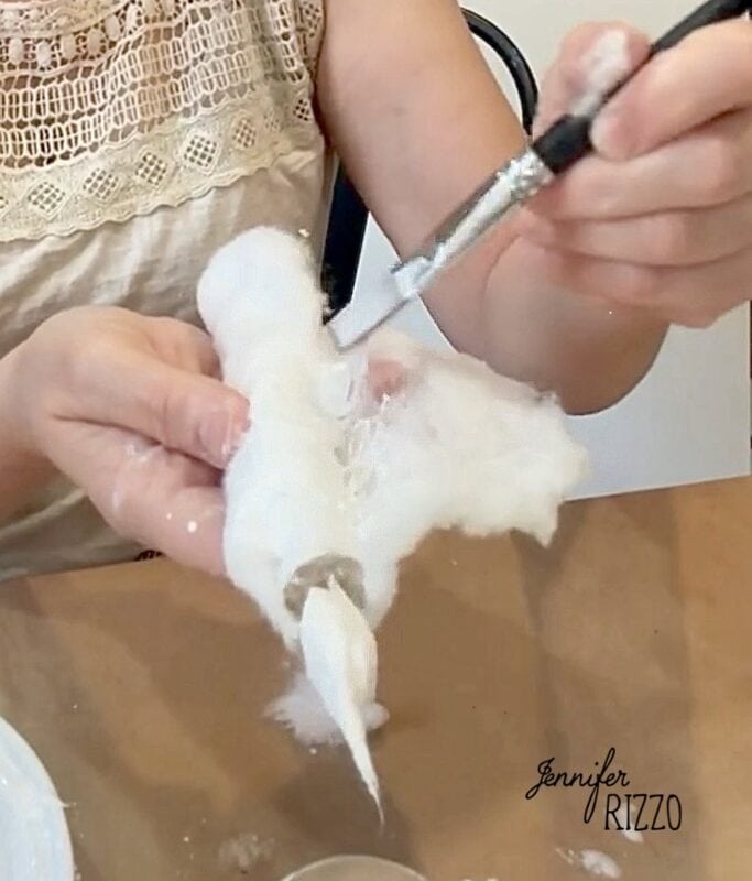Person sculpting a small figure with white material using a metal tool at a table. The name “Jennifer Rizzo” appears on the lower right corner of the image.