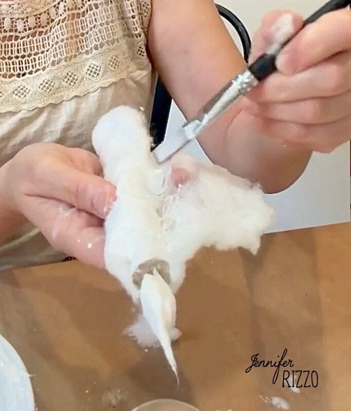 Person sculpting a small figure with white material using a metal tool at a table. The name “Jennifer Rizzo” appears on the lower right corner of the image.