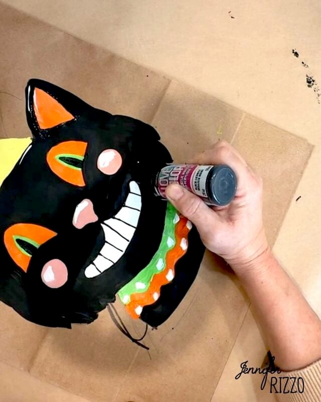 A hand decorates a painted black cat face with a party hat, orange and green collar, and a wide grin using a bottle of white paint on brown craft paper.