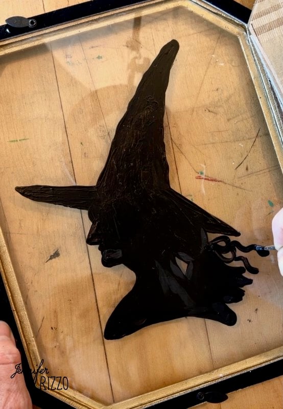 A person paints a black silhouette of a witch with a pointed hat on a glass panel, with wood visible underneath.