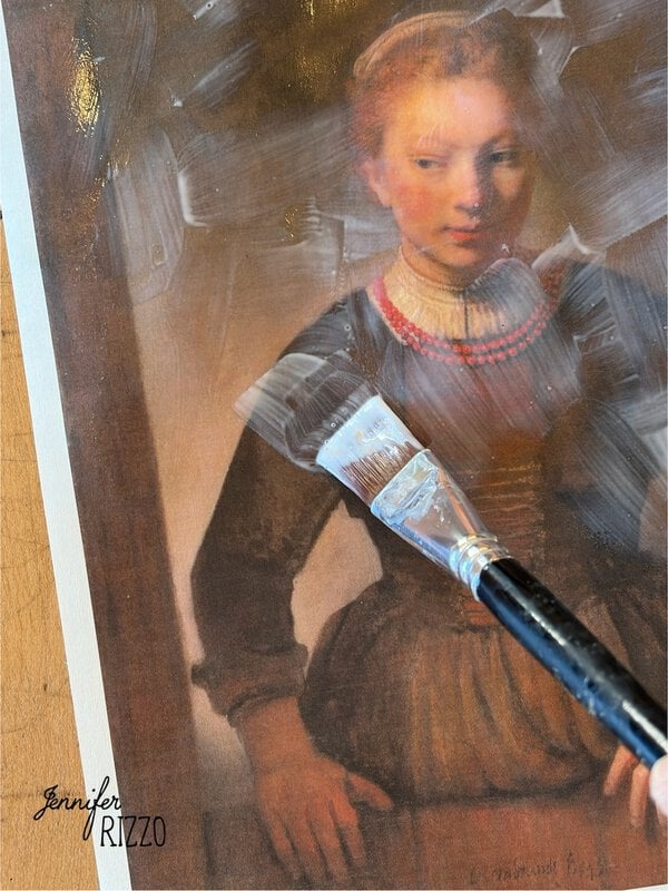 A paintbrush applies white brushstrokes over a printed portrait of a woman in historical dress on a wooden surface.