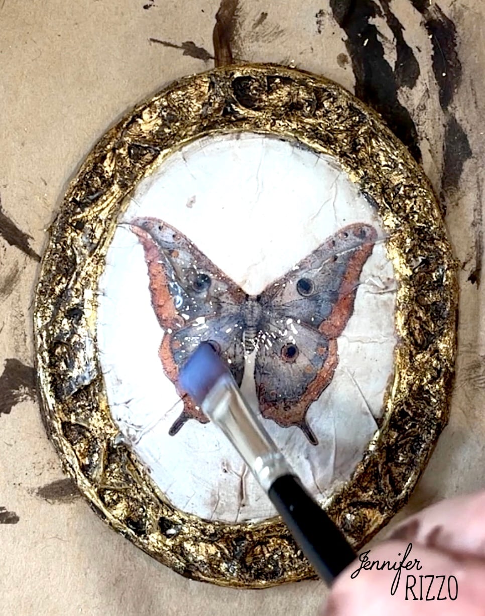 A hand uses a brush to apply medium to a circular artwork featuring a butterfly, bordered by a textured, gold-painted frame.