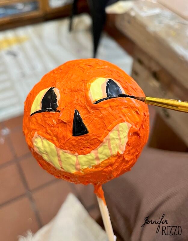 A hand paints the eye of an orange papier-mâché pumpkin face on a stick. The pumpkin has a wide yellow grin and triangular nose. “Jennifer Rizzo” is written in the corner.
