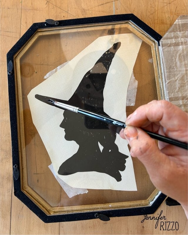 A hand uses a paintbrush to outline a black witch silhouette on glass, with a paper template underneath.