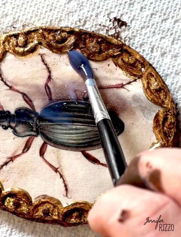 A hand uses a paintbrush to add a blue glaze to the gold border of an oval artwork featuring a beetle on a textured surface.
