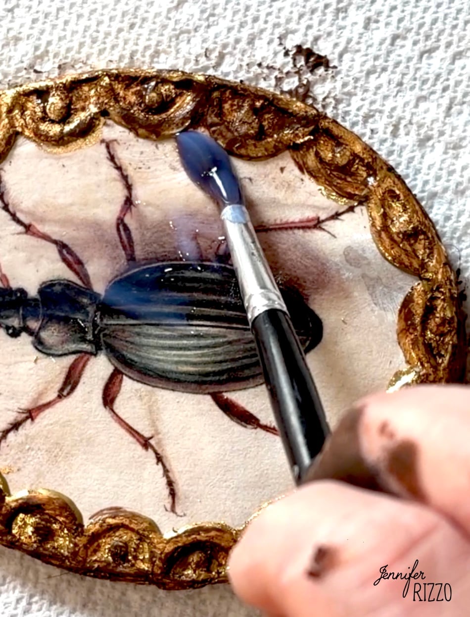 A hand uses a paintbrush to add a blue glaze to the gold border of an oval artwork featuring a beetle on a textured surface.