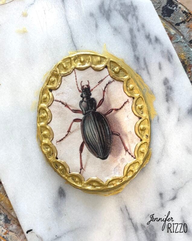 A round air dry clay decorated with a detailed beetle illustration and a gold ornate border sits on a marble surface for Air Dry Clay Faux Insect Taxidermy