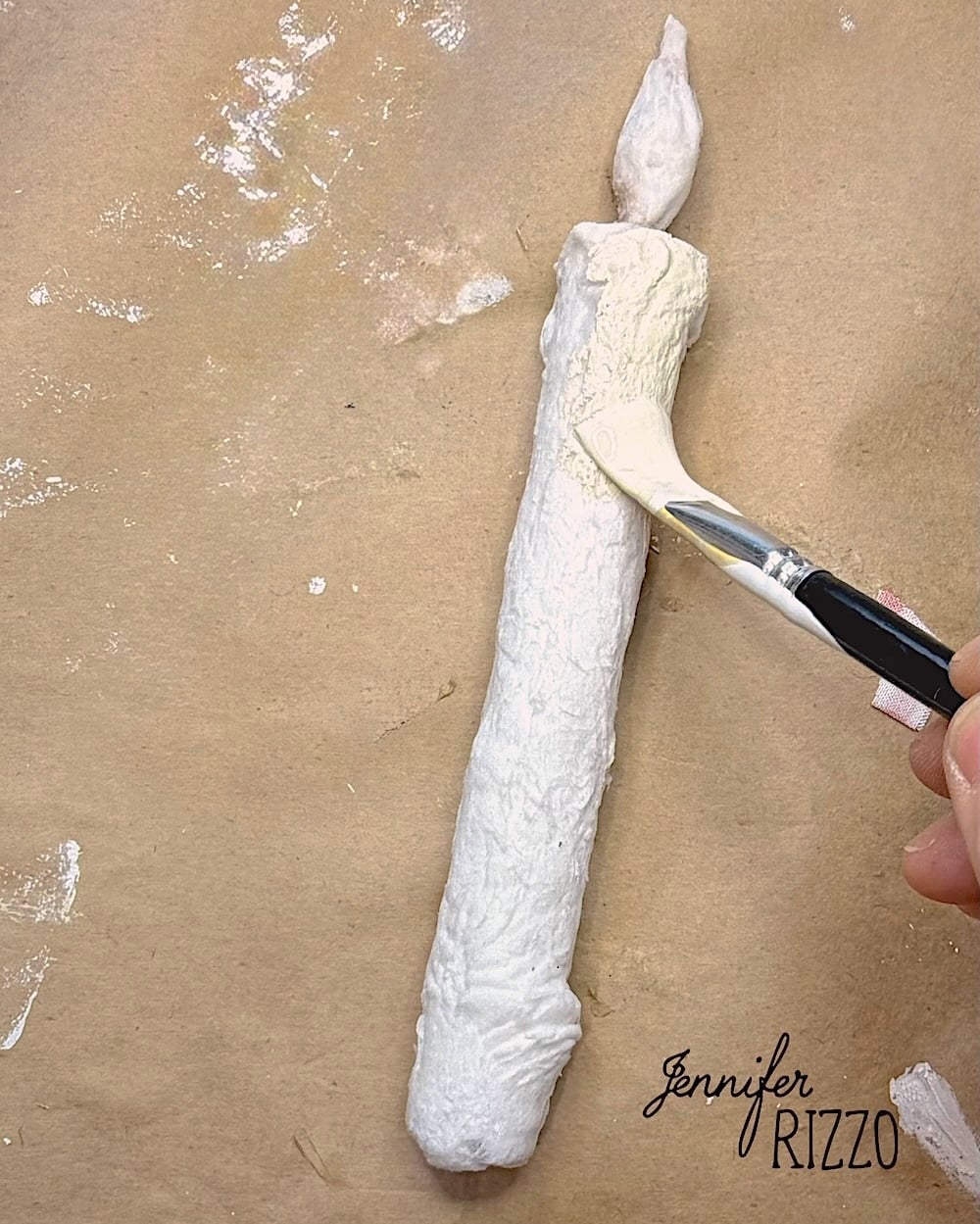 A hand paints a textured, white sculpted candle with a flame on a brown surface, reminiscent of Spun Cotton Faux Candles. The name “Jennifer Rizzo” appears in the lower right corner.