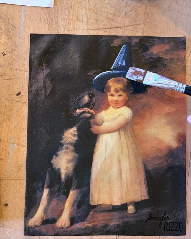A hand paints a black witch hat onto a vintage portrait of a child in white dress standing next to a large black and white dog.