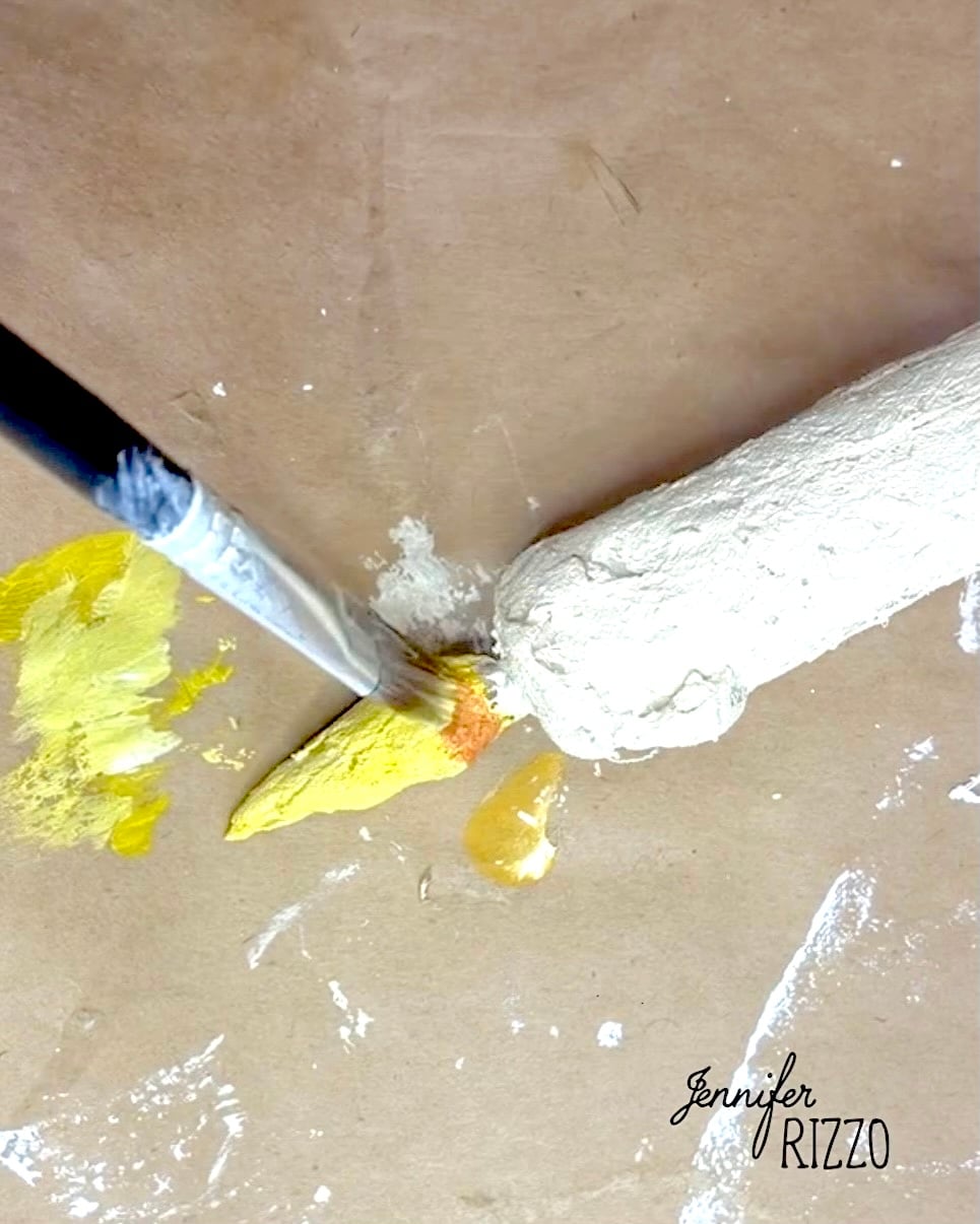A paintbrush applies yellow paint to the beak of a textured, white bird sculpture on a brown surface.