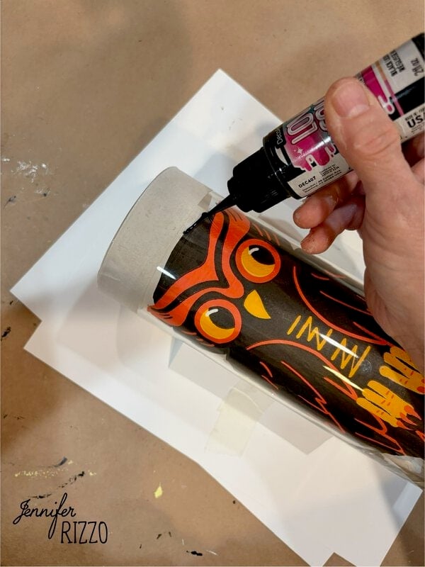 A hand uses black paint to outline an orange owl design on a tall glass vase, which is taped to a white paper.