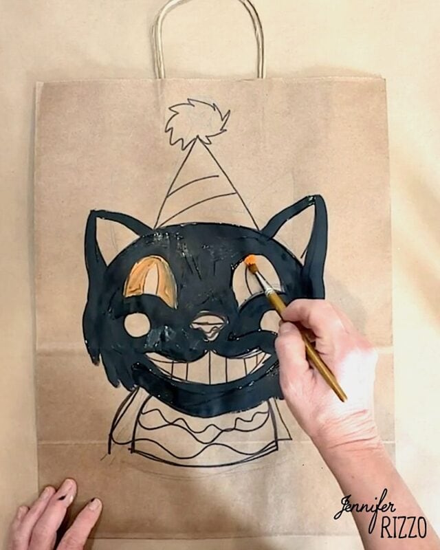 A hand paints a black cat face with a party hat on a brown paper bag using a small paintbrush.
