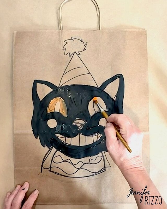 A hand paints a black cat face with a party hat on a brown paper bag using a small paintbrush.