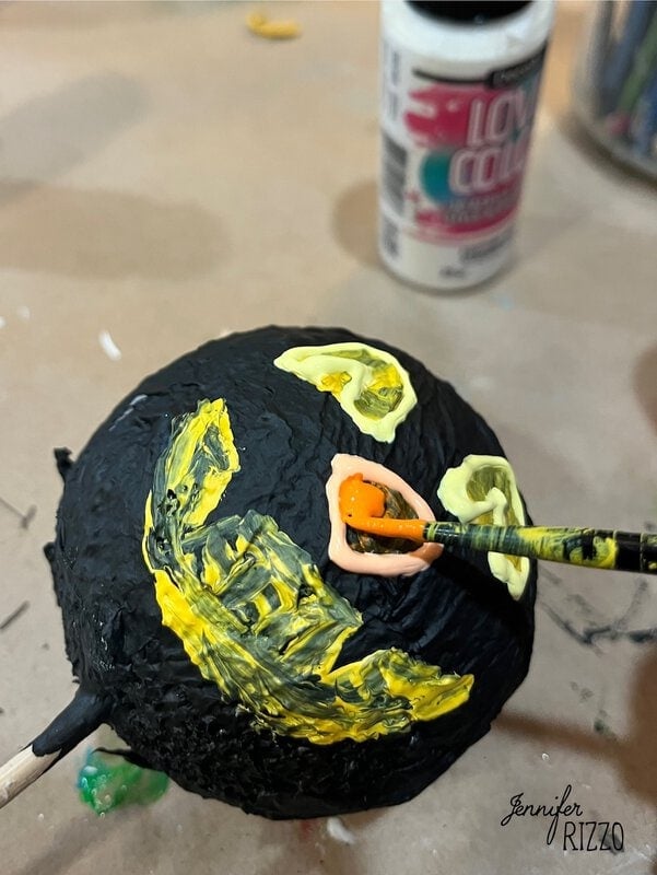 A hand paints yellow and orange features on a black round object, possibly a Halloween decoration, with paint bottles and a stick visible on a brown surface.