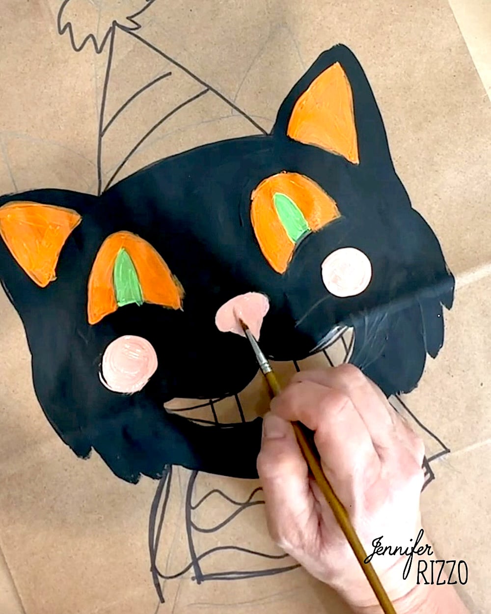 A hand paints a black cat face with orange and green details on brown paper. The cat wears a party hat, and the artist’s signature “Jennifer Rizzo” appears in the corner.