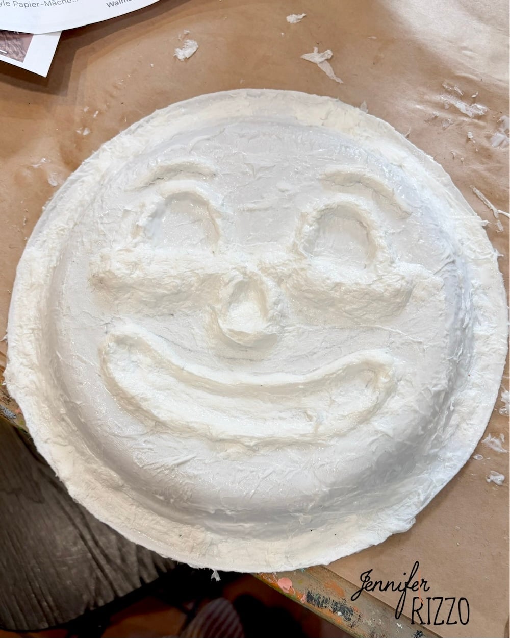 A paper mache plate molded into a smiling face, with raised eyes, eyebrows, and mouth, on a brown surface.