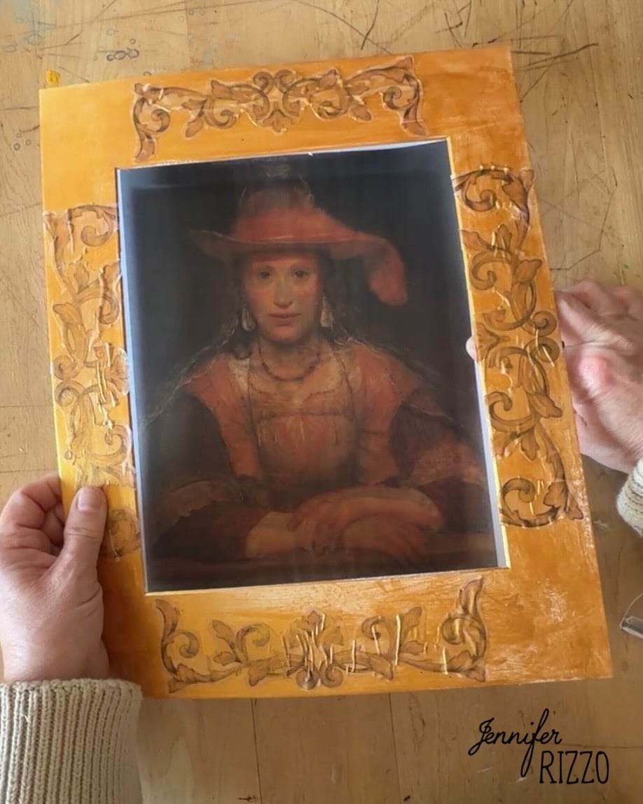 A person holds a framed print of a classical portrait with an ornate, hand-decorated yellow border on a wooden table.