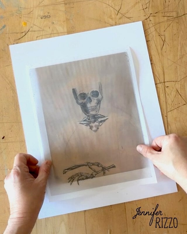 Two hands hold a translucent paper with a skeleton drawing over a white sheet on a wooden table. “Jennifer Rizzo” is written in the bottom right corner.